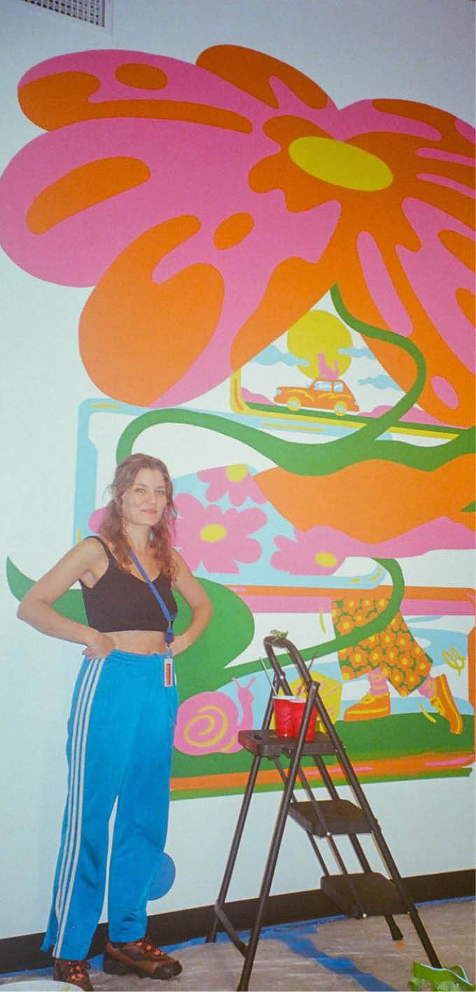 Two adults stand in front of a large colorful mural featuring a stylized flower and abstract shapes while a small step ladder with a red cup on it is positioned between them. Description generated by AI